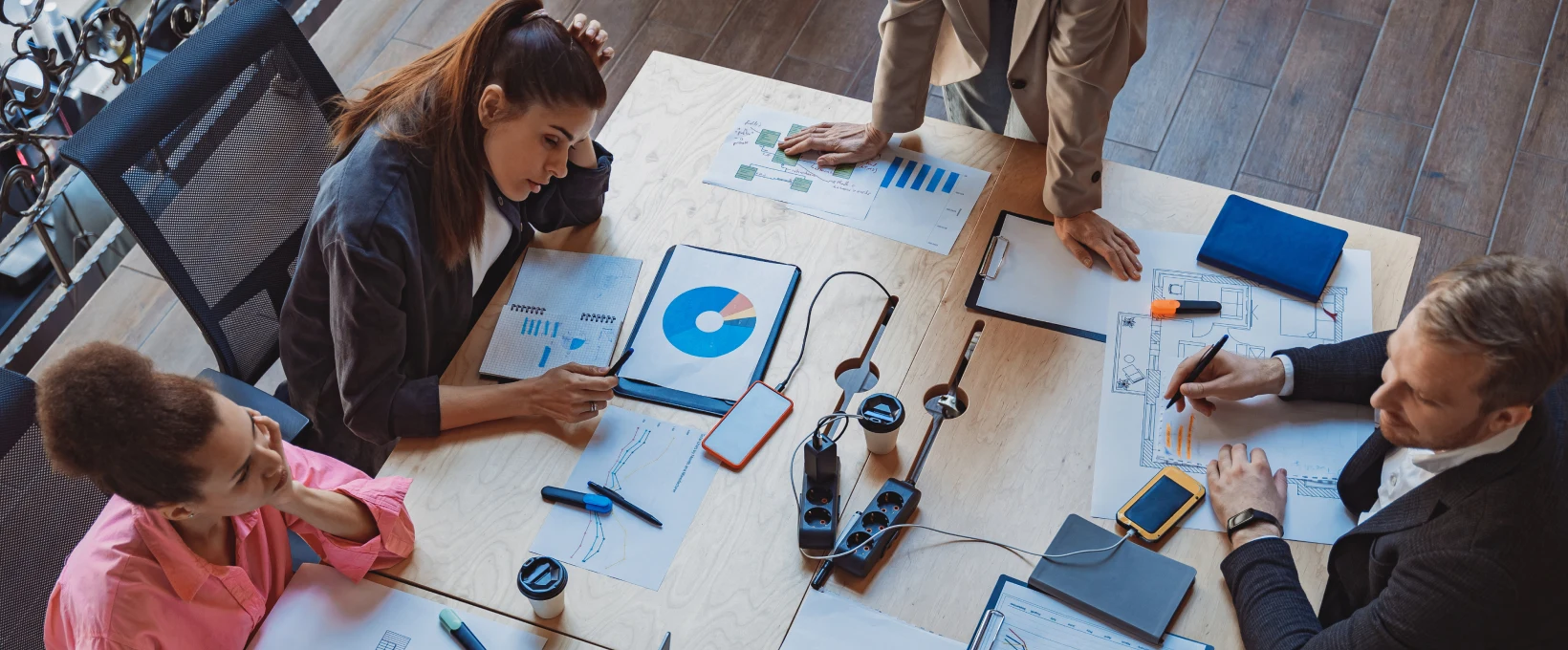 Overhead view of four people gathered around a conference table during a business meeting. Charts with pie and line graphs, a floor plan, clipboards, notebooks, coffee cups, smartphones, and power strips are spread across the table while the group reviews documents and discusses plans.